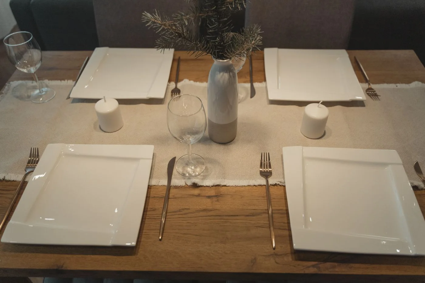 A wooden table set with square white plates, pale napkins, cutlery and glasses.