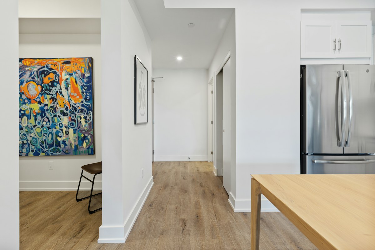 A pale hallway beside a kitchen with timber flooring, a colorful picture and a refrigerator.