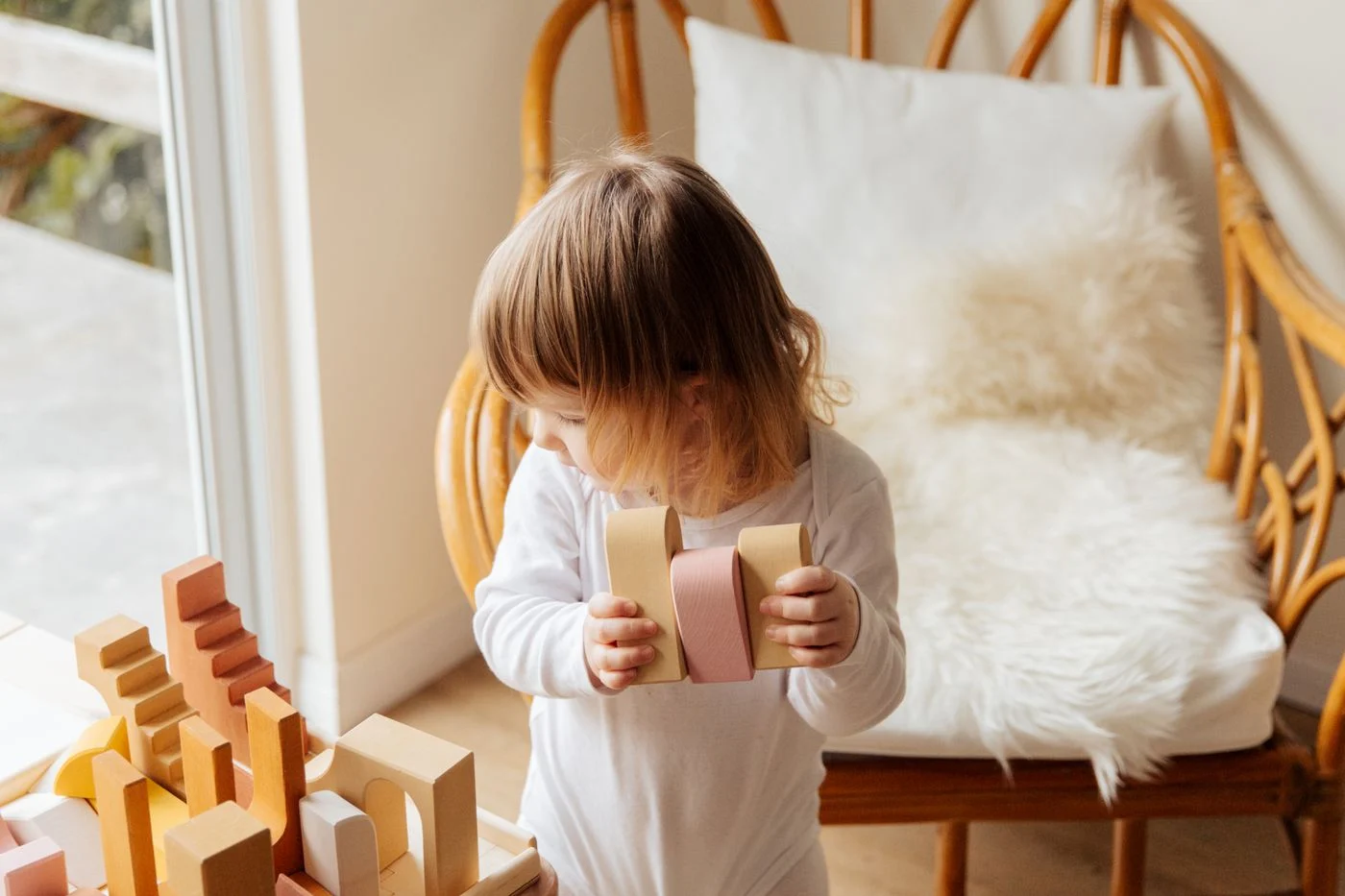Child holding wooden building blocks beside a wicker chair