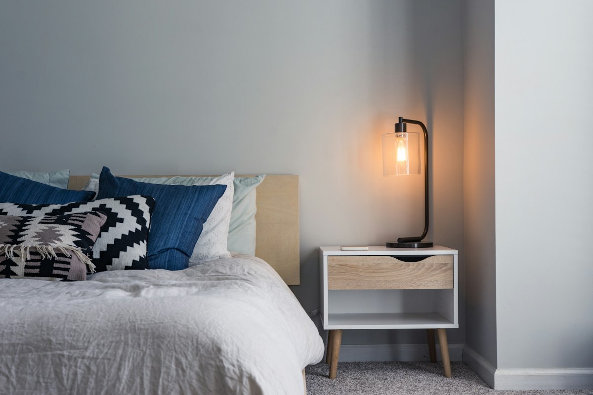 A bed with blue cushions beside a pale cabinet and a lit lamp