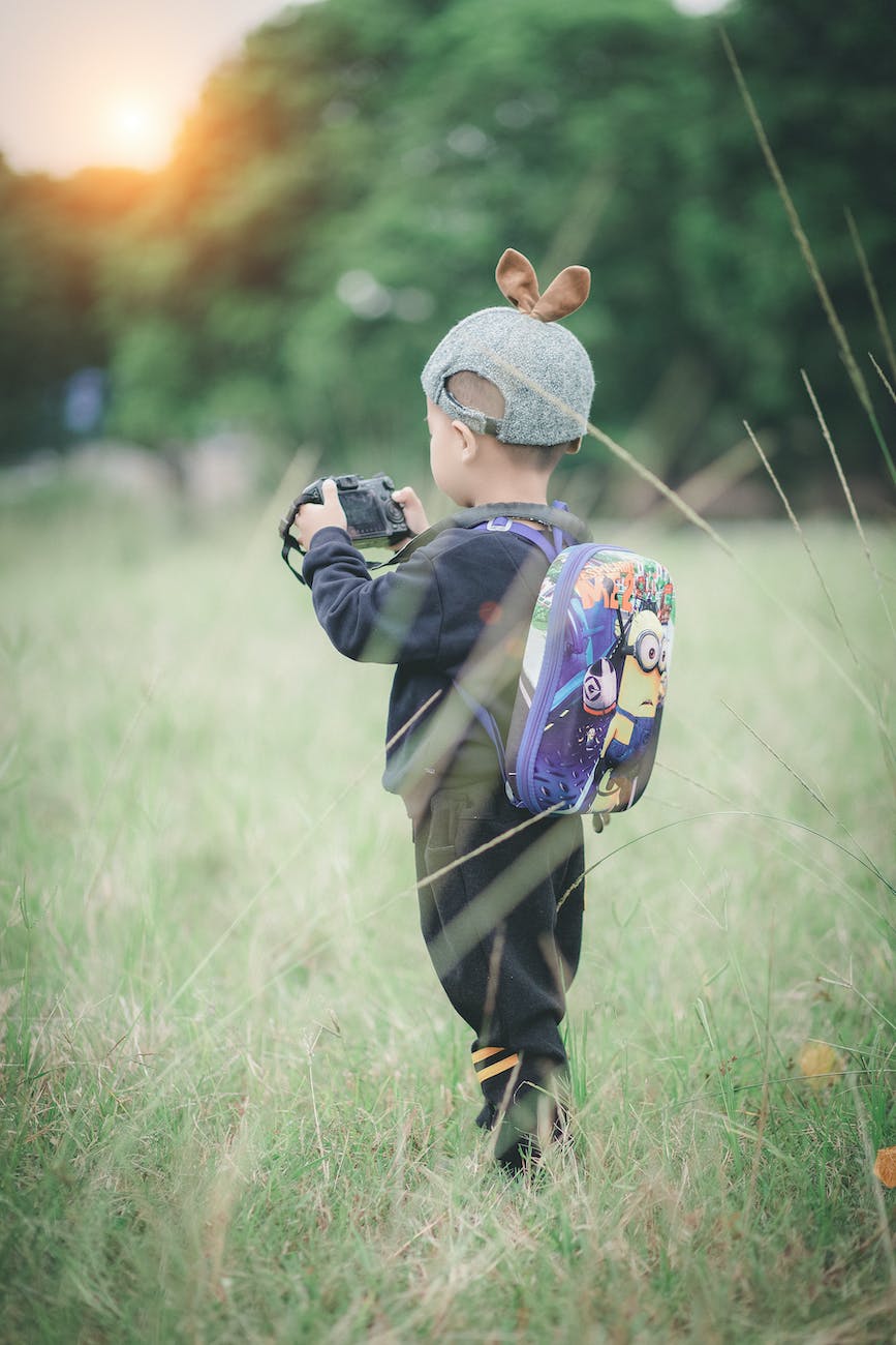 boy carrying backpack holding camera standing on green grass