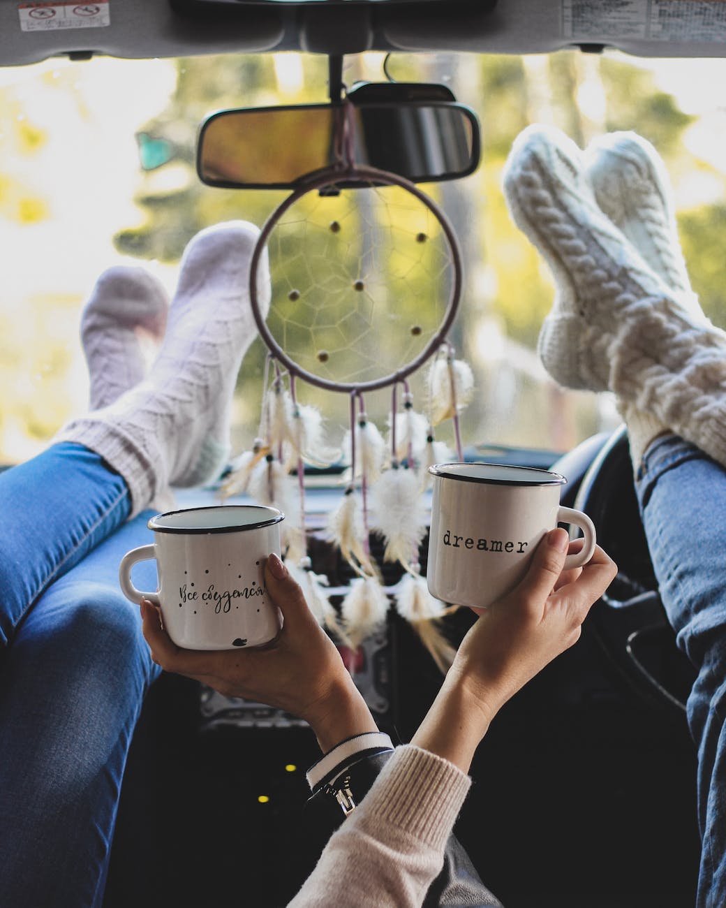 two people holding coffee mugs with their feet on the dashboard of a vehicle with an ornament hanging on the rear view mirror