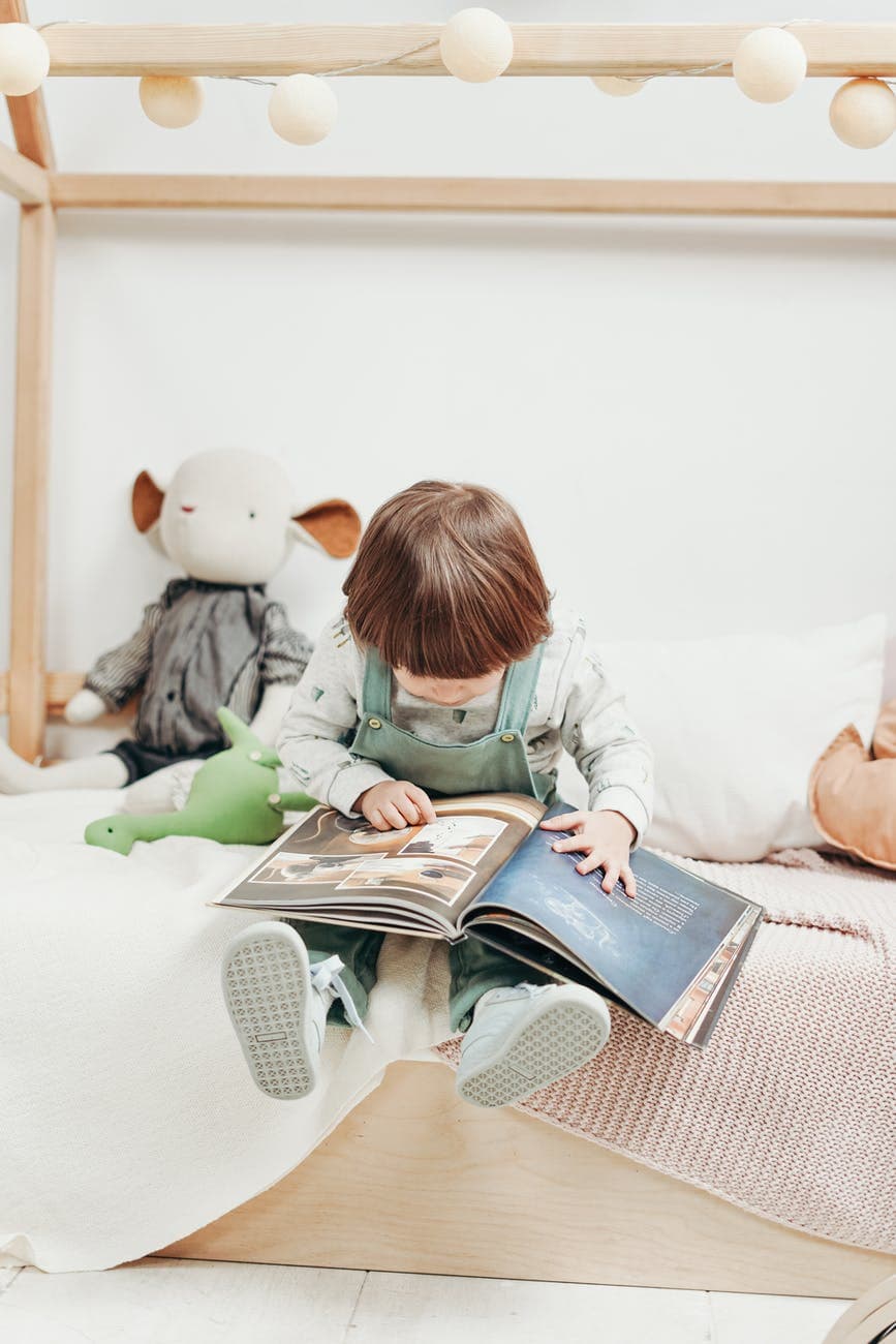 child in white long sleeve top and dungaree trousers sitting on bed reading book