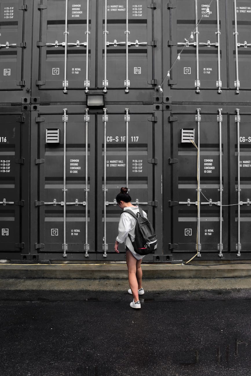 photography of woman in gray long sleeve shirt carrying black backpack standing in front of black intermodal container